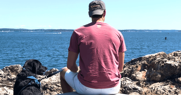 Person wearing a 3Ways North Shore Beef hat sitting on the rocks by the ocean with a black dog, looking out at the water on the Massachusetts North Shore.
