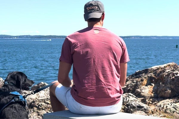 Person wearing a 3Ways North Shore Beef hat sitting on the rocks by the ocean with a black dog, looking out at the water on the Massachusetts North Shore.